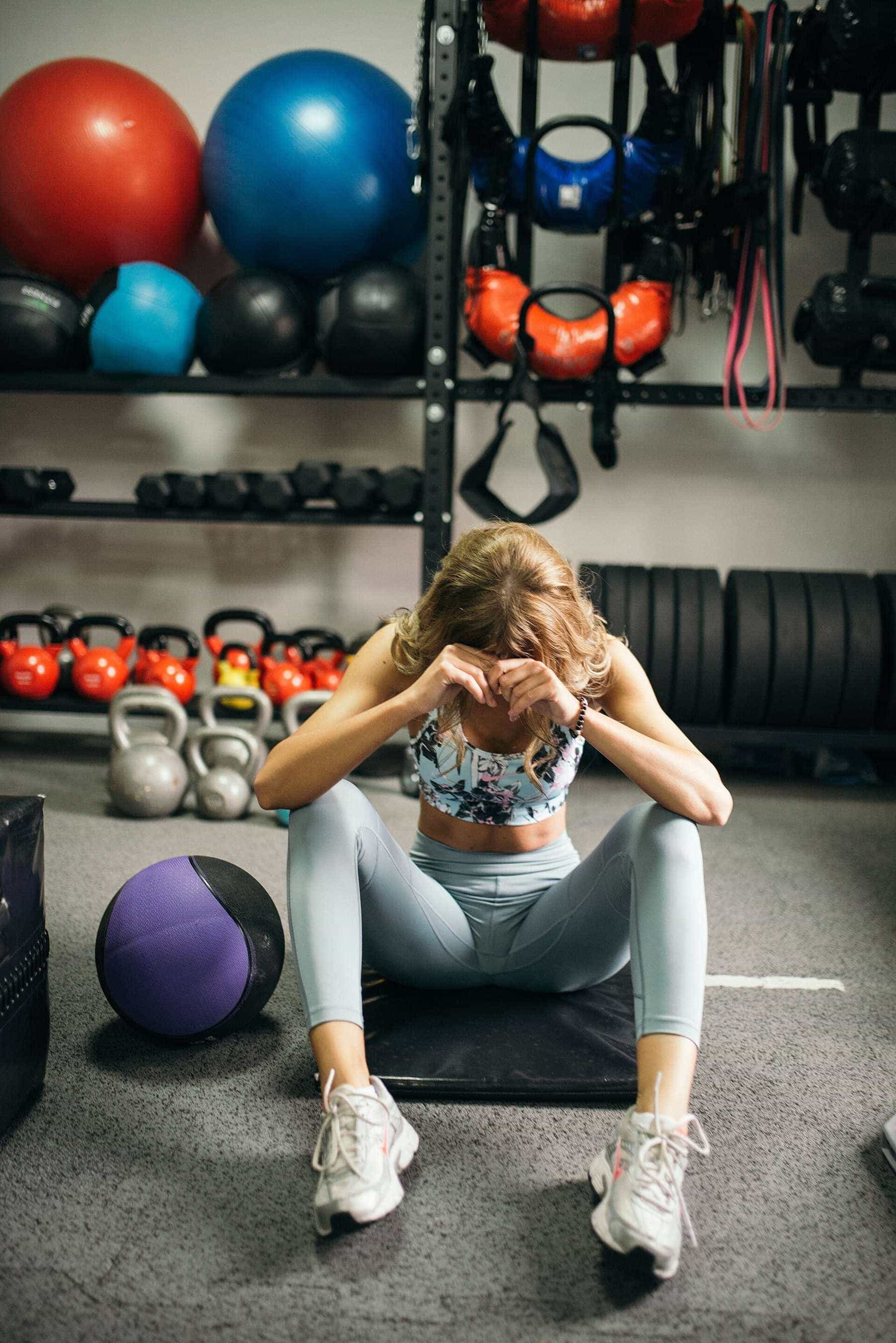 A woman sitting on a gym floor with her head in her hands after a workout