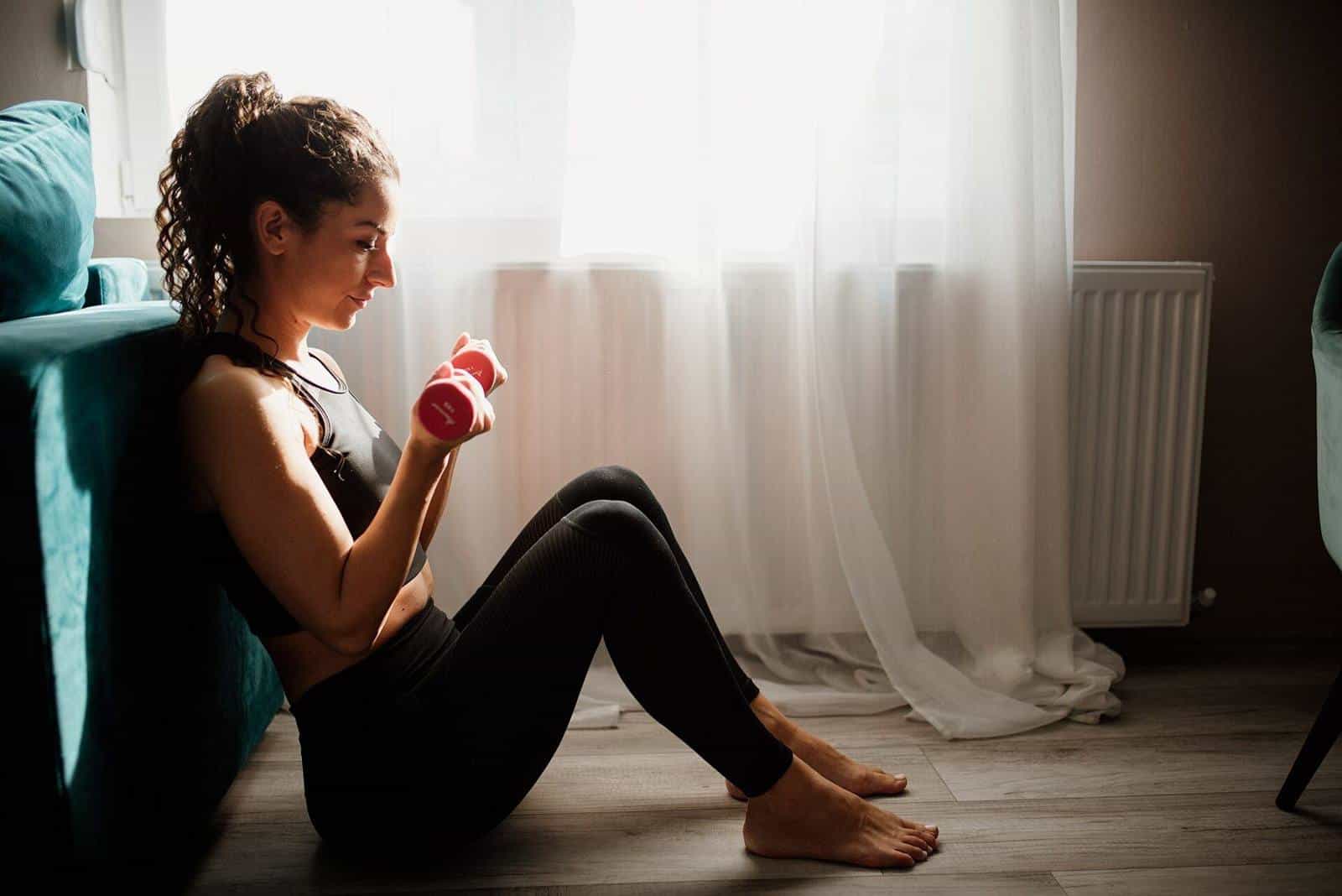 A woman sitting on the floor by a sunny window lifting a pink dumbbell