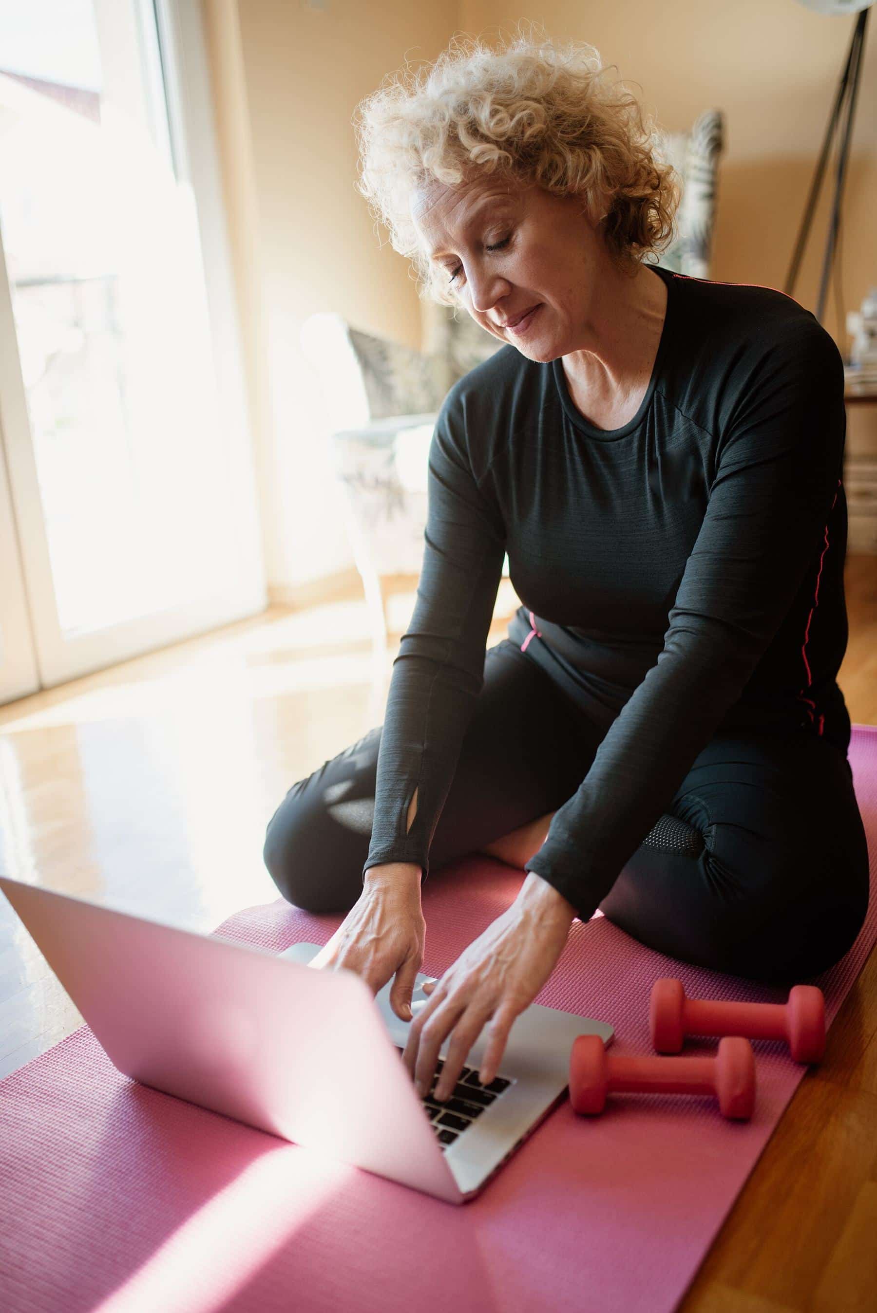 An older woman on a pink mat with dumbbells, following a workout on a laptop