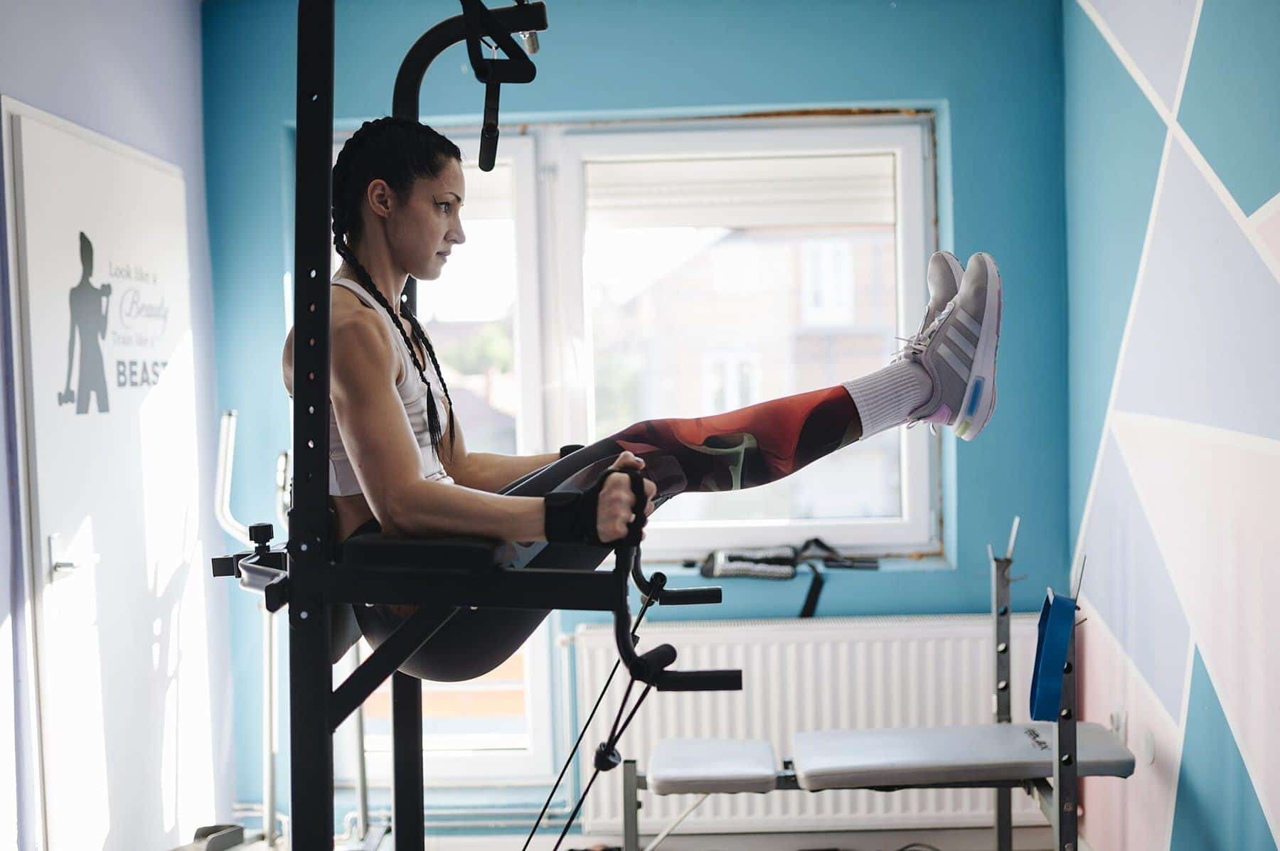 A woman doing knee raises on a dip station beside a bright window at home